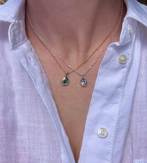 Two Birthstone Droplet Pendants worn layered with a white linen shirt: bronze with an emerald on a gold filled chain, and sterling silver with an alexandrite