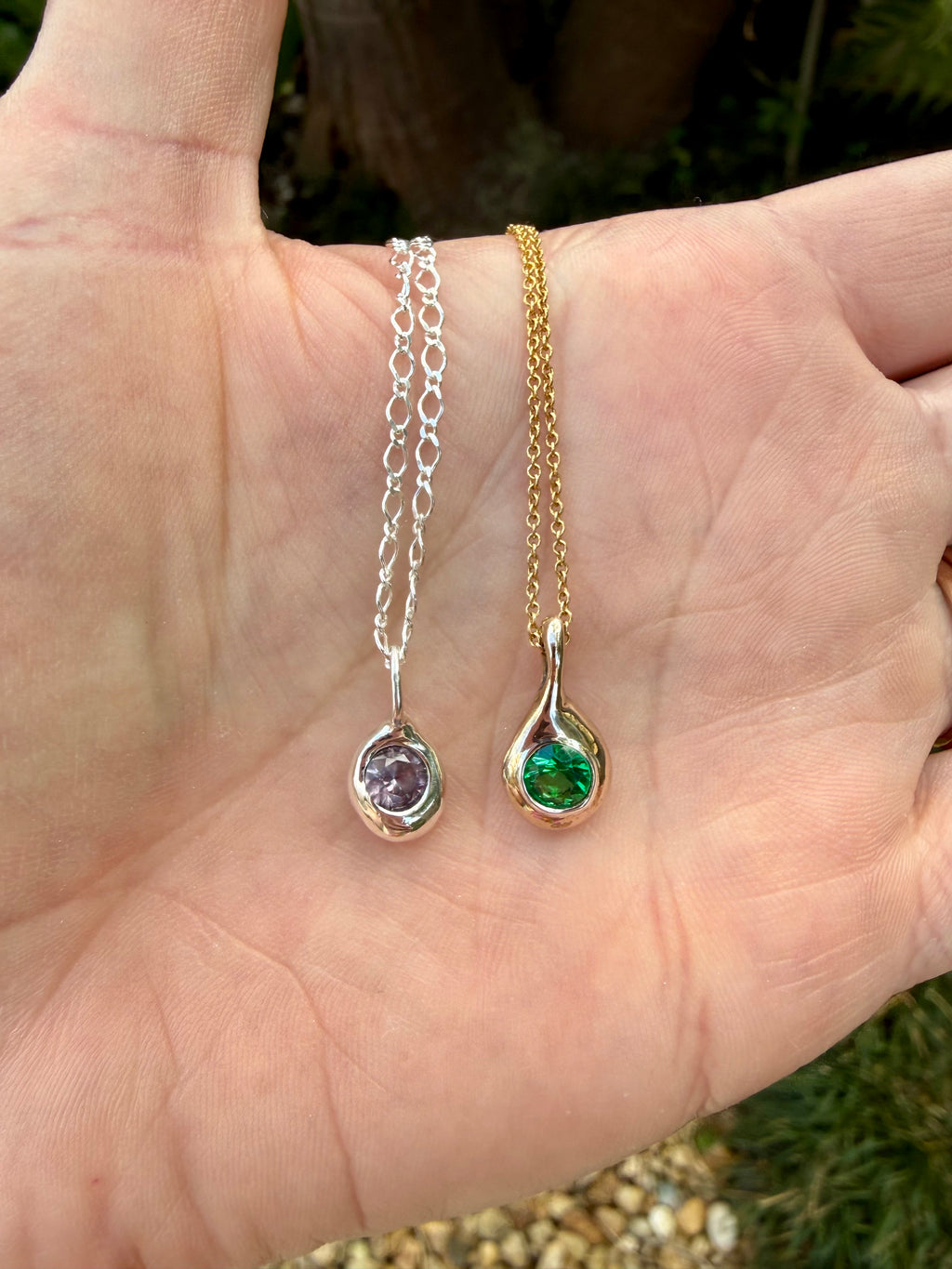 Birthstone Droplet Pendants held in an open palm: sterling silver with an alexandrite and bronze with an emerald on a gold filled chain