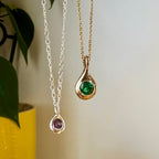 Birthstone Droplet Pendants hanging beside a plant: bronze with an emerald on a gold filled chain and sterling silver with an alexandrite