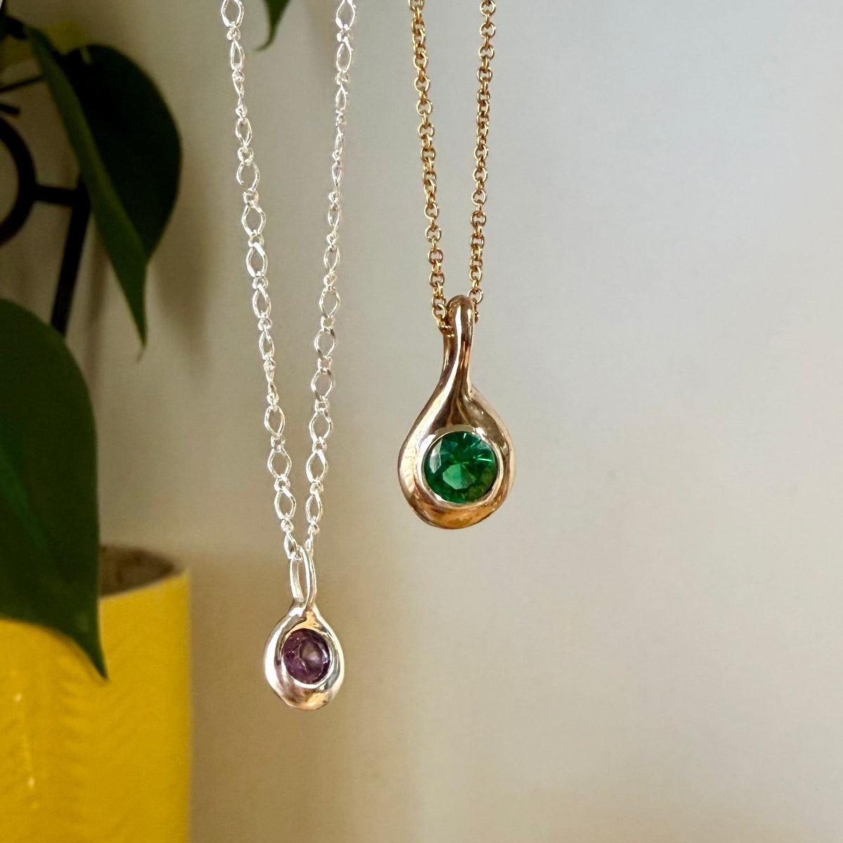 Birthstone Droplet Pendants hanging beside a plant: bronze with an emerald on a gold filled chain and sterling silver with an alexandrite