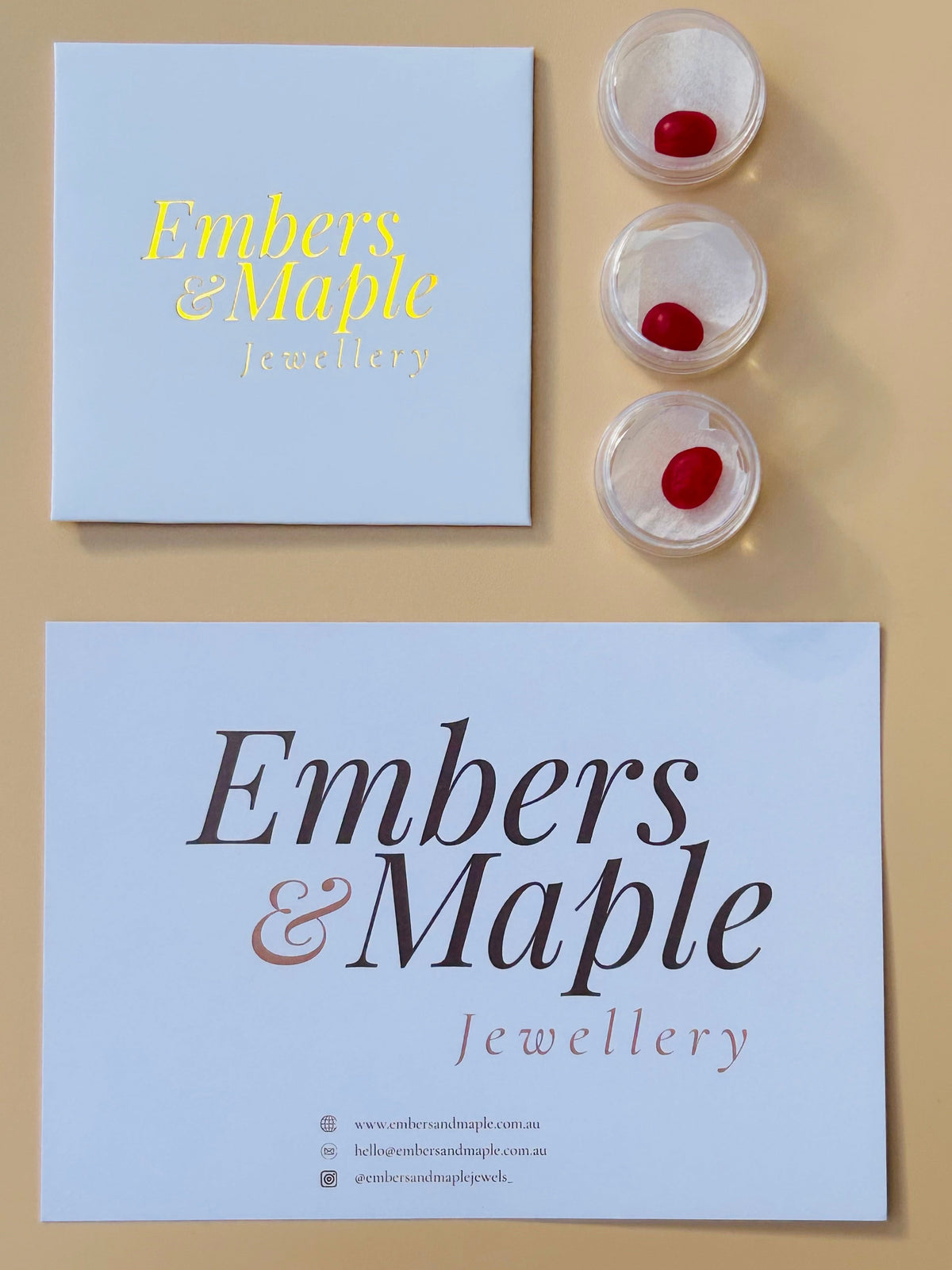 A styled flat-lay of a jewellery box and a matching branded card on a beige background, alongside three small clear round containers holding red jewellers wax. 